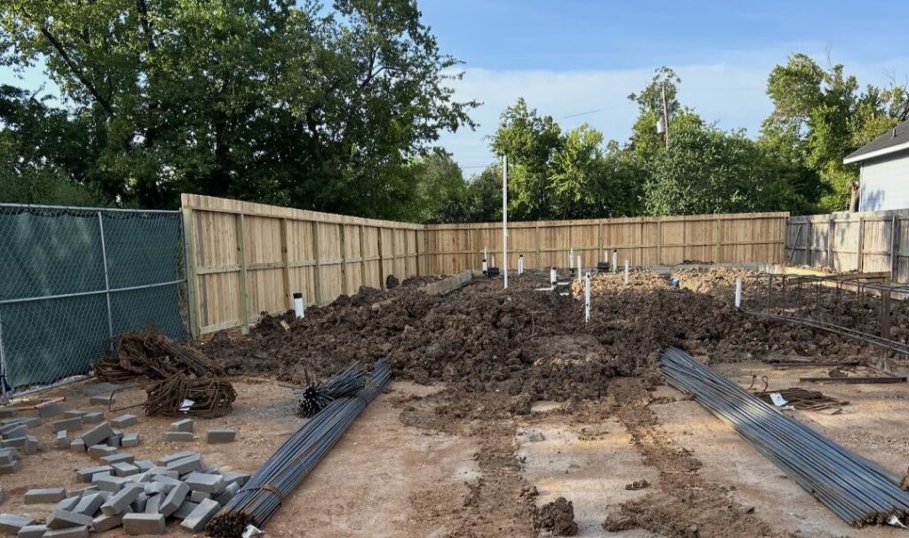 Cleared construction site at Boy Street with mounded soil, bundled rebar, stacked pavers, and white plumbing pipes, surrounded by a new wooden privacy fence and trees.