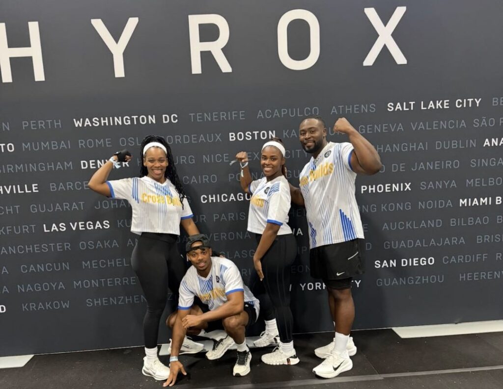 Four team members in matching white jerseys smile and flex in front of a HYROX event backdrop.